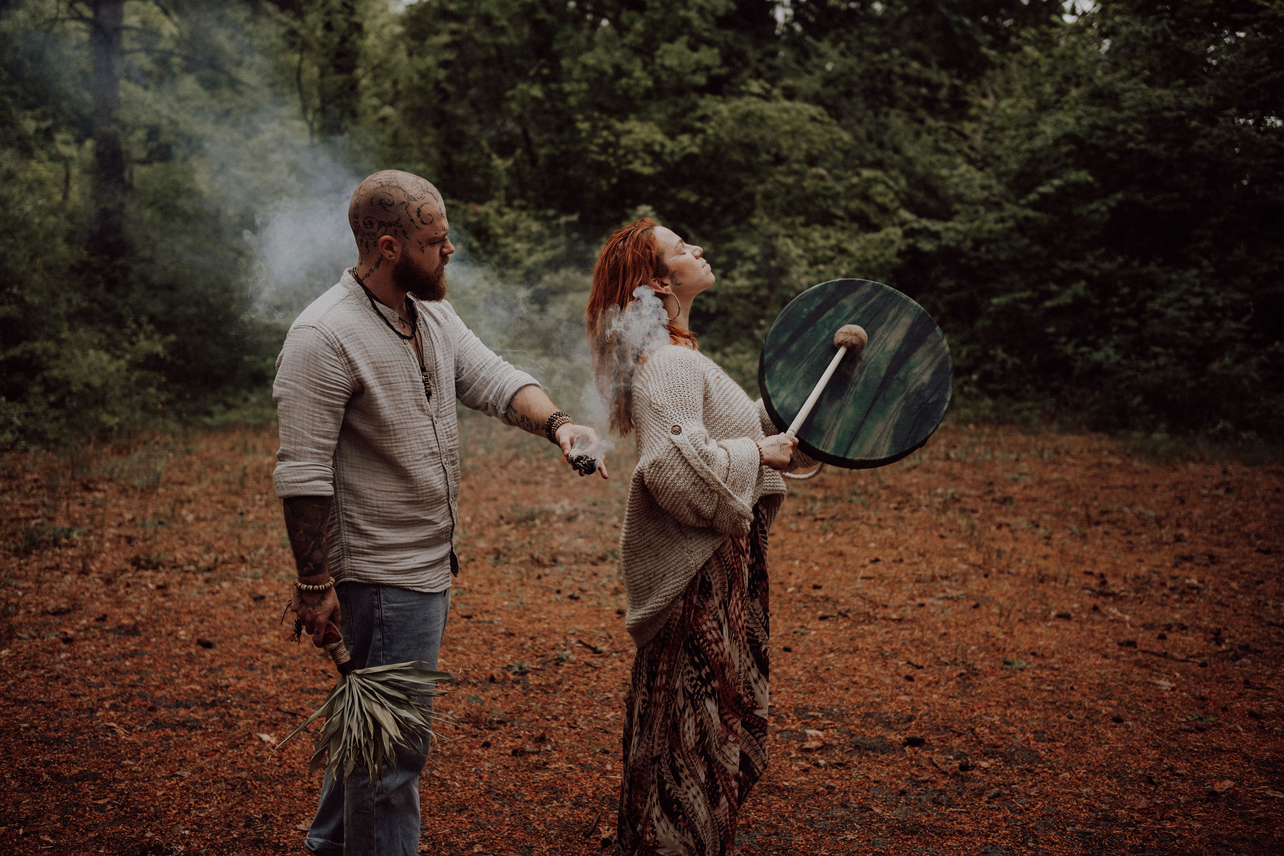 Rituel en forêt : un homme fait passer une fumée autour d'une femme rousse, yeux fermés, tambour chamanique à la main