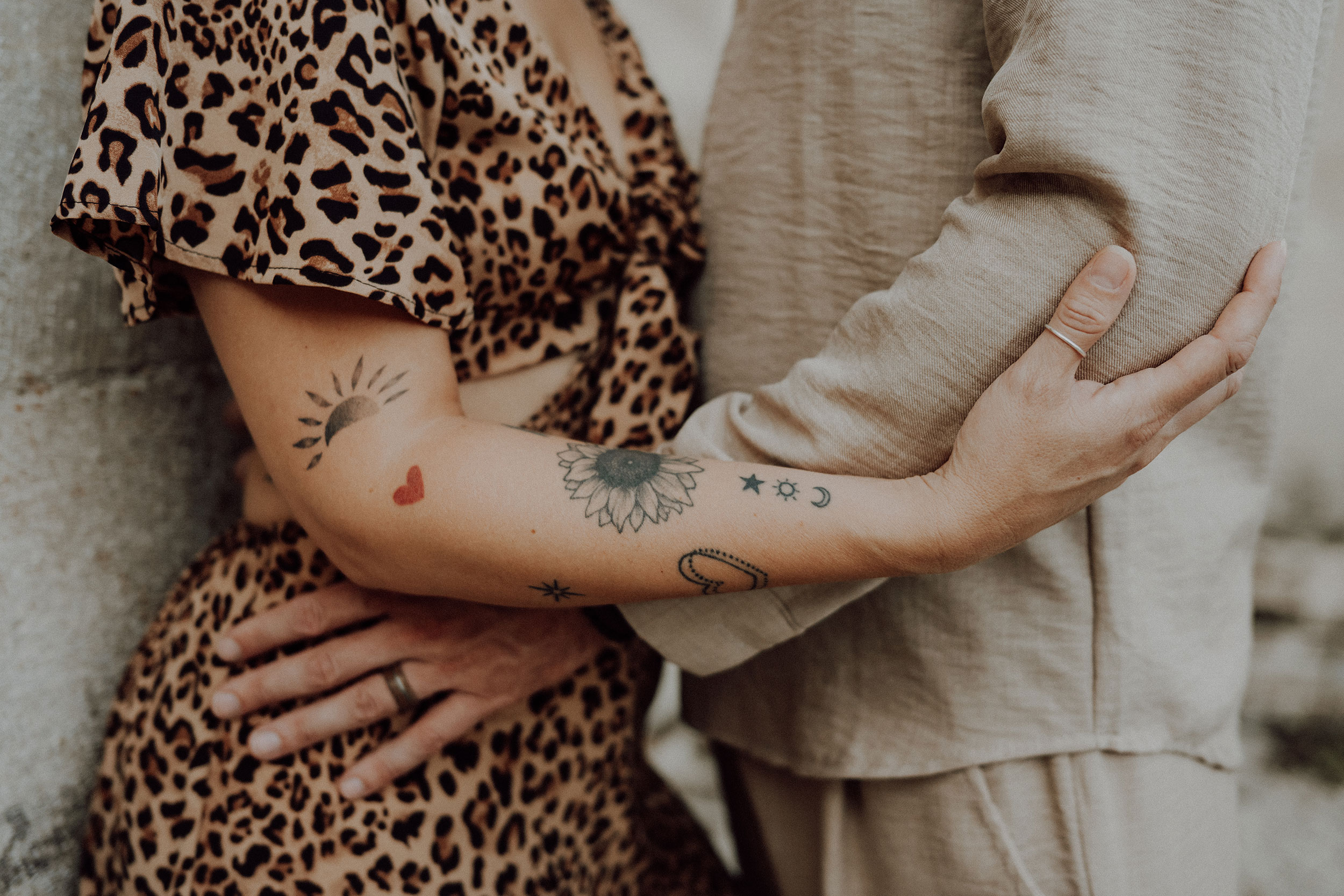 Bras enlacés en gros plan : avant-bras tatoué d'un tournesol et d'un soleil, main de l'homme posée sur la robe léopard