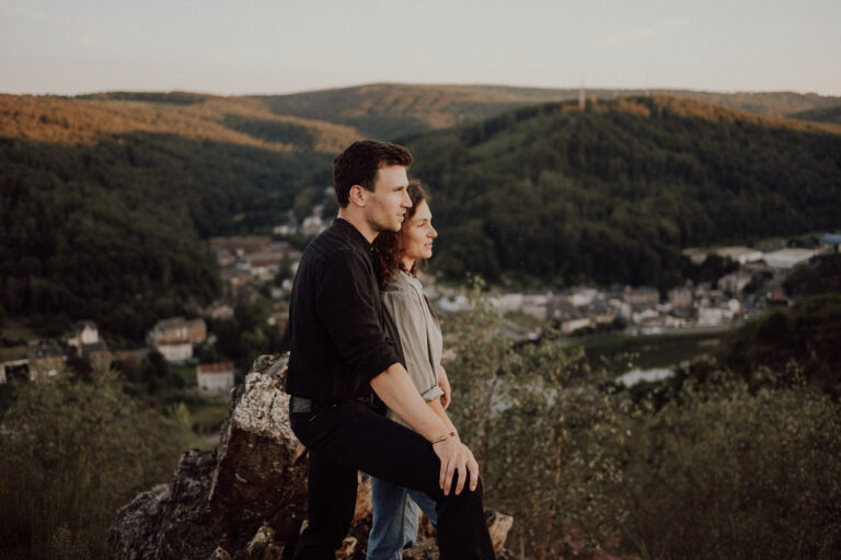 Couple serré l'un contre l'autre sur un rocher, le regard tourné vers une vallée boisée et la ville en contrebas, en fin de journée