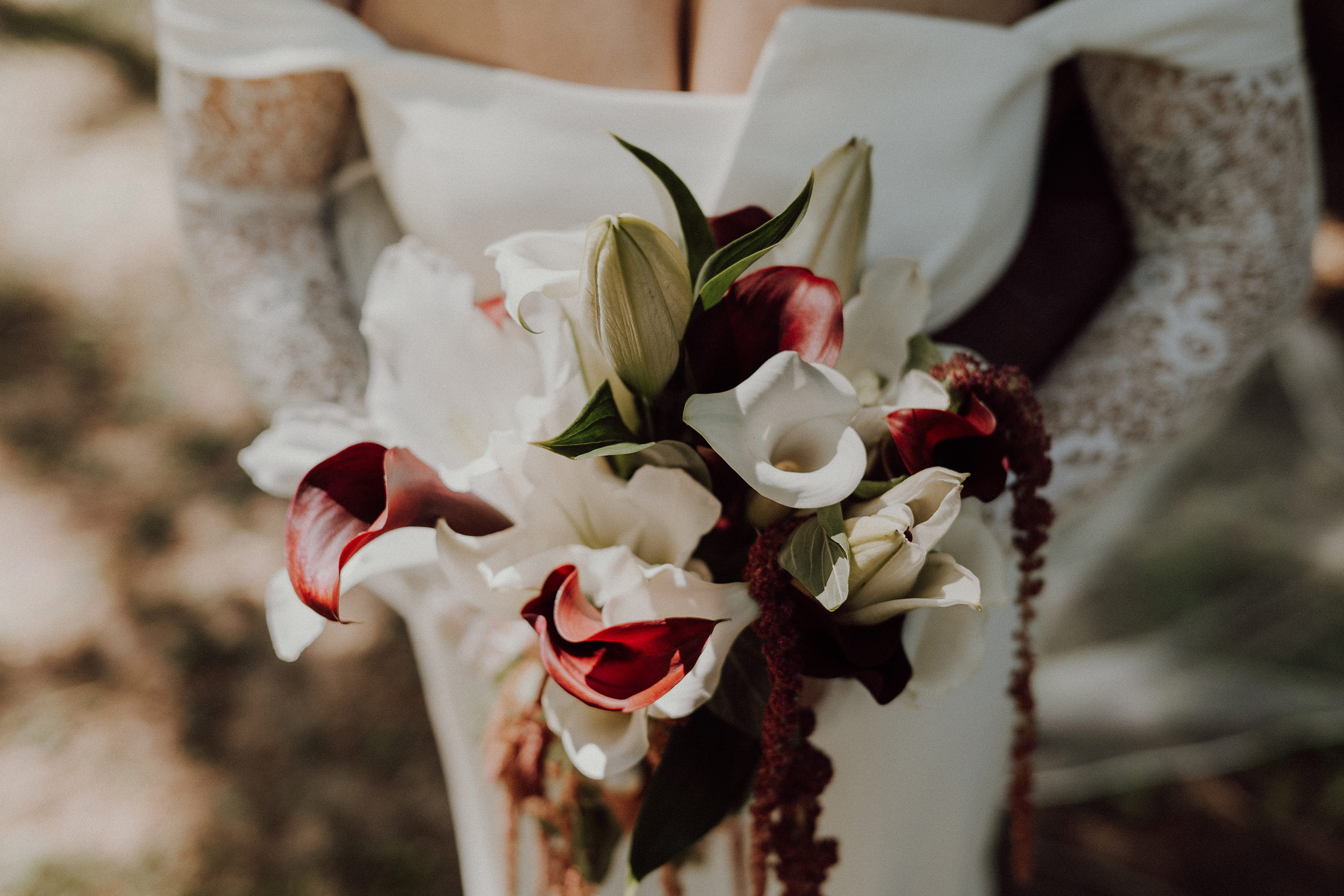 Gros plan sur le bouquet de la mariée, lys blancs, arums bordeaux et amarante retombante, devant sa robe à manches de dentelle