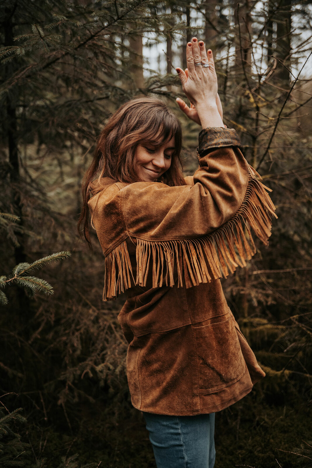 Camille, photographe Eyes of Cam, sourit les yeux baissés, bras levés au-dessus de la tête, dans un sous-bois de conifères