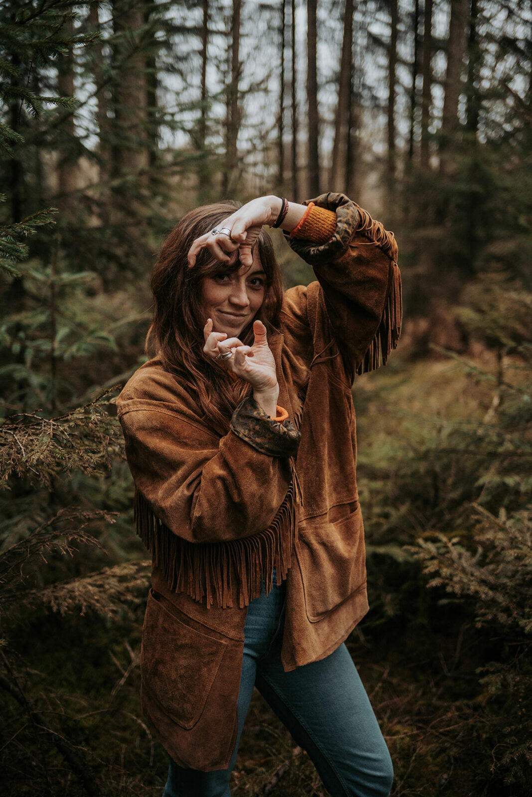 Camille, photographe Eyes of Cam, mime un appareil photo avec ses mains, en veste en daim à franges dans un sous-bois de conifères