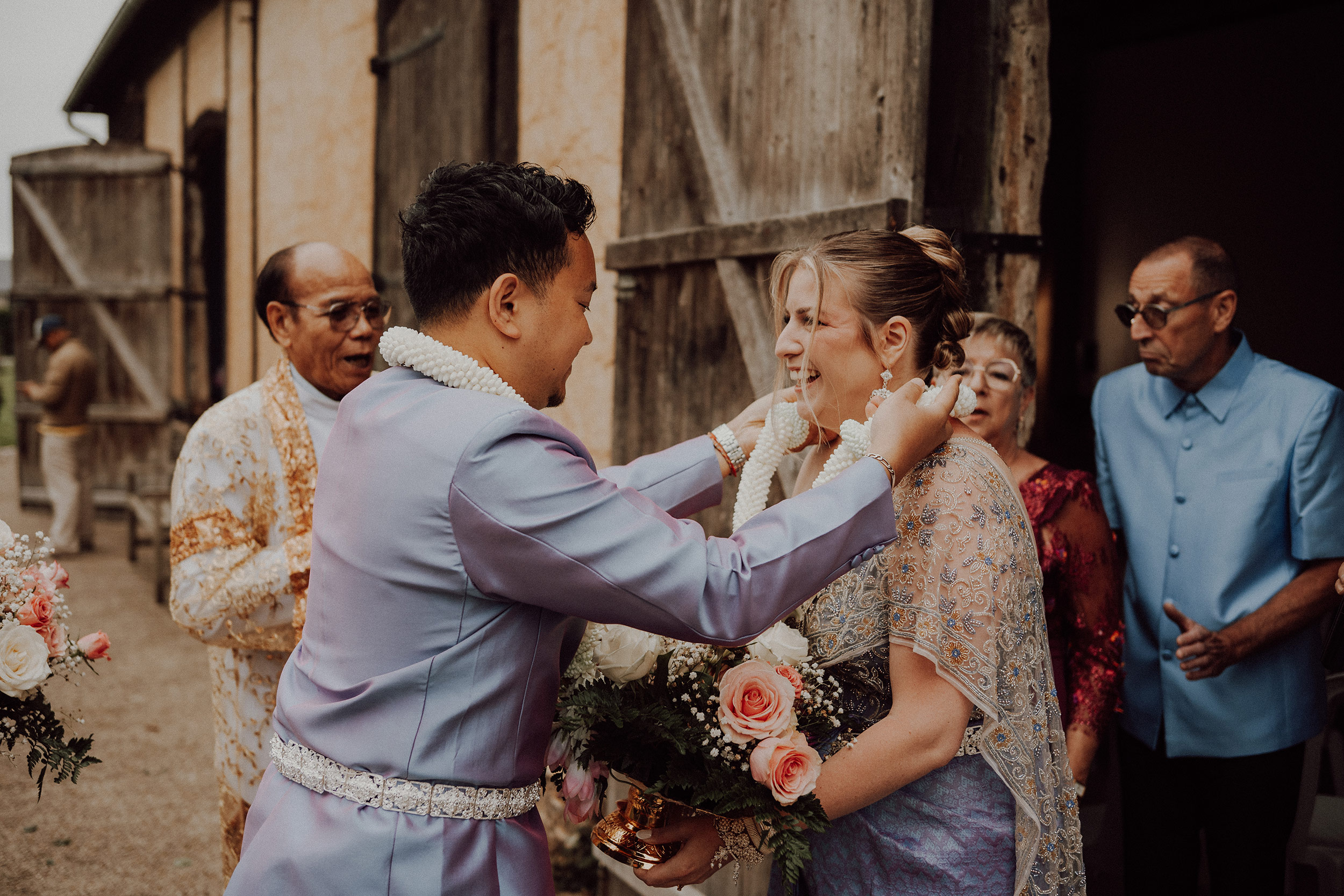 Marié en tenue traditionnelle lilas qui passe un collier de fleurs à la mariée, entourés de leur famille devant une grange