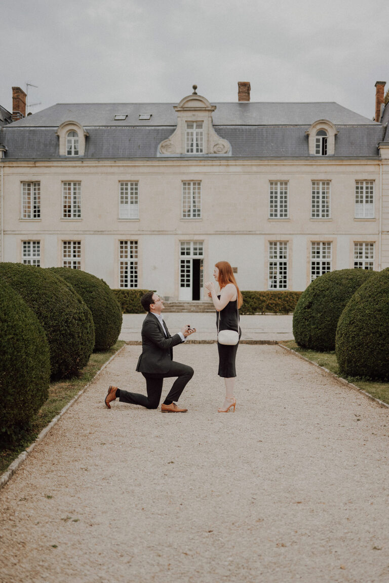 Demande en mariage : un homme à genou tend une bague à sa compagne rousse, devant la façade d'un château en pierre