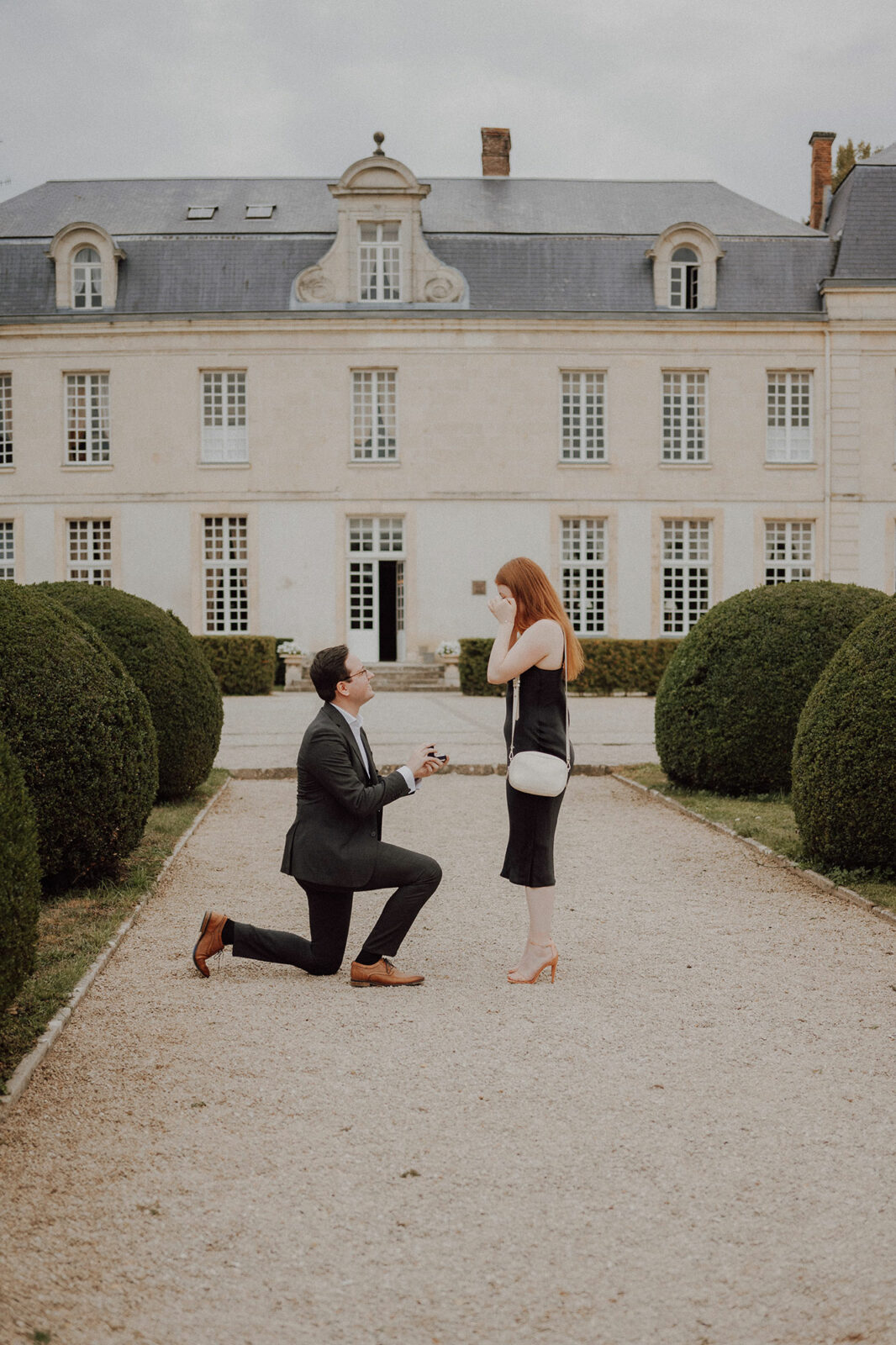 Demande en mariage : la jeune femme porte la main à son visage, émue, face à son compagnon à genou dans l'allée d'un château