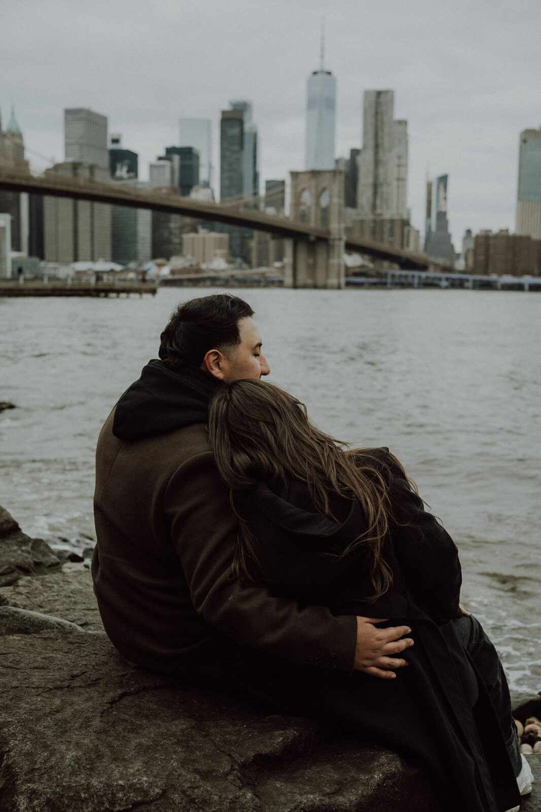 Fiancés assis de dos sur un rocher, elle la tête sur son épaule, face au pont de Brooklyn et à Manhattan