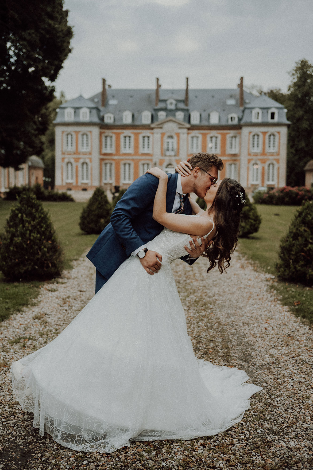 Baiser renversé des mariés sur l'allée de gravier d'un château en briques et pierre, bordée d'arbustes taillés en cône
