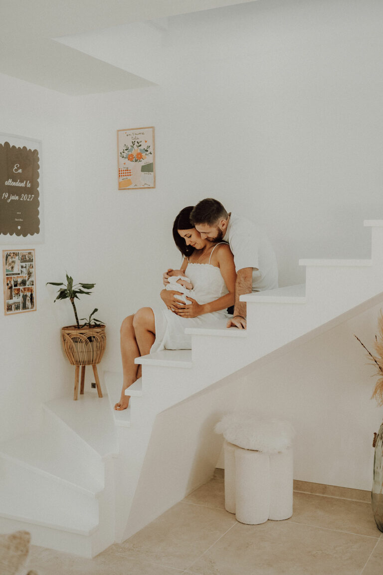 Jeunes parents assis sur un escalier blanc avec leur nouveau-né dans les bras, chez eux
