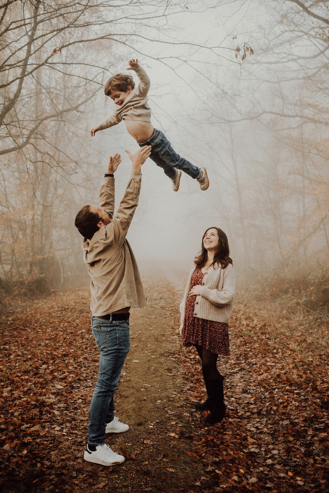 Papa qui lance son fils en l'air sous le regard de la future maman, dans une allée de forêt brumeuse