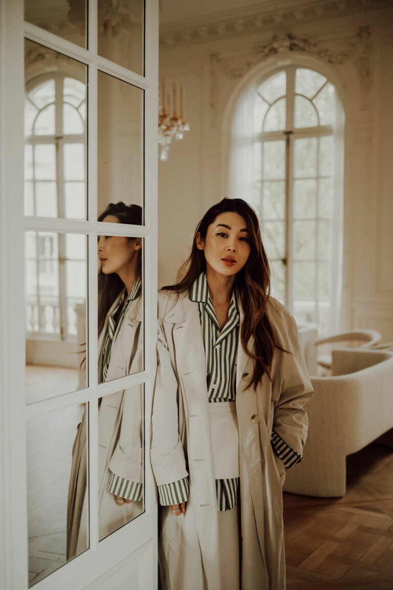 Femme en trench beige et chemise rayée appuyée contre une porte-fenêtre, son reflet dans la vitre, dans un salon à moulures