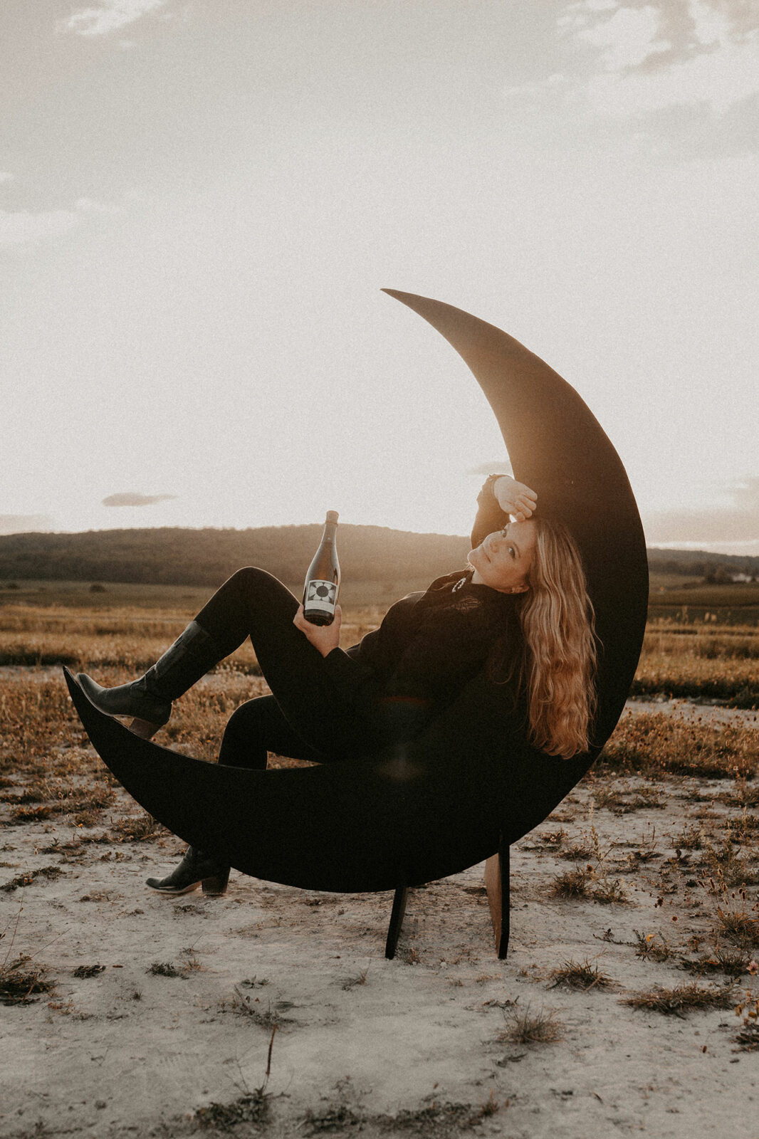 Femme blonde en noir allongée dans un croissant de lune géant, bouteille à la main, sur une terre crayeuse au coucher du soleil