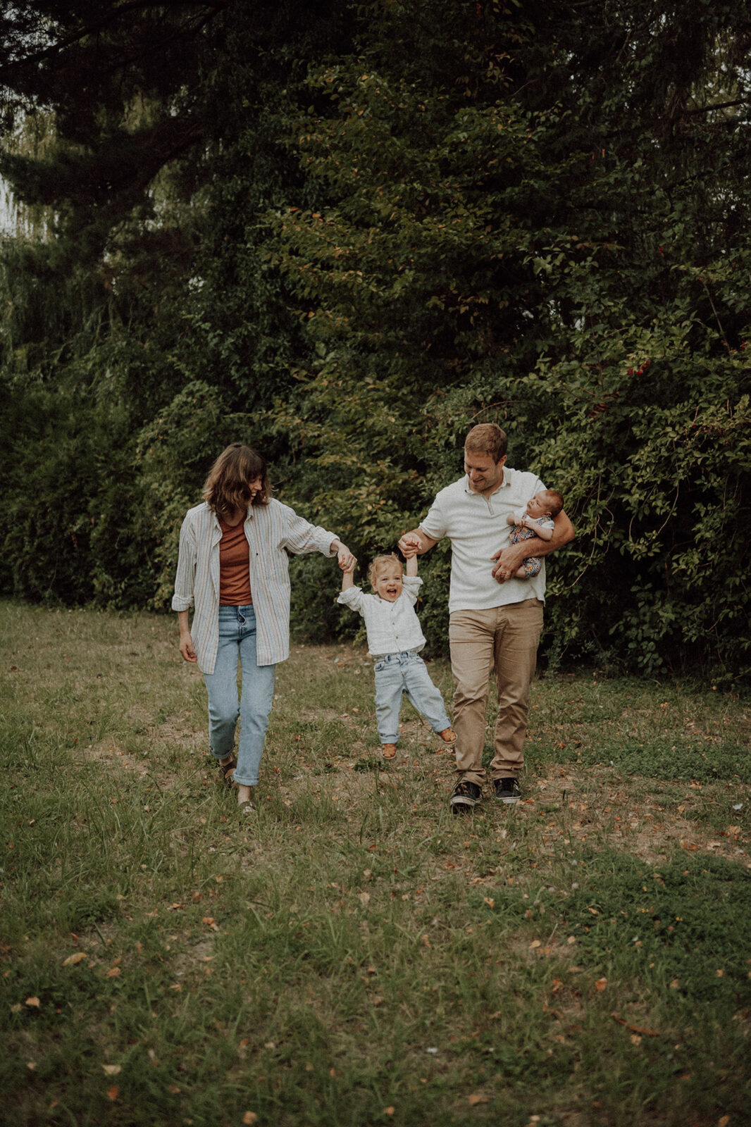 Parents qui balancent leur enfant hilare en le tenant par les mains, le papa porte un nouveau-né au creux du bras, dans un pré bordé d'arbres